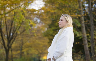 Eine blonde Frau mit weißem Mantel läuft durch einen herbstlichen Wald und atmet tief ein.