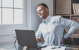 Ein Mann an einem Schreibtisch vor einem Computer, hinter ihm steht ein Regal. Er fasst sich mit schmerzverzerrtem Gesicht an den Rücken.
