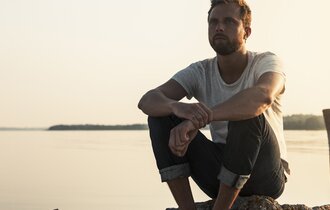 Mann sitzt am Wasser auf Steinen und schaut in die Ferne.