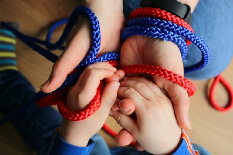 Nahaufnahme der Hände eines Kindes und eines Erwachsenen, die beide in Strickseile eingewickelt sind.