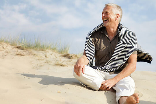 Ein Mann gehobeneren Alters, der auf dem Sand einer Düne sitzt. Über ihm leicht bewölkter Himmel, neben ihm Schilfgras.