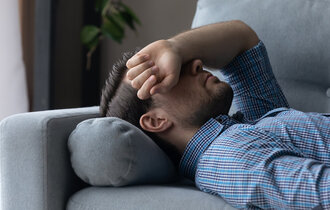 Ein Mann mit kariertem Hemd liegt auf einem hellblauen Sofa und hat einen Arm über seine Augen gelegt.