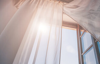  Ein geöffnetes Fenster, der Vorhang ist zur Seite gezogen und die Sonne scheint herein.