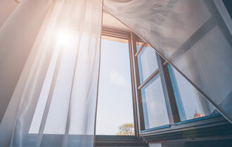  Ein geöffnetes Fenster, der Vorhang ist zur Seite gezogen und die Sonne scheint herein.