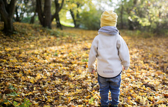 Ein Kleinkind läuft mit Mütze durch das Laub eines Waldes.