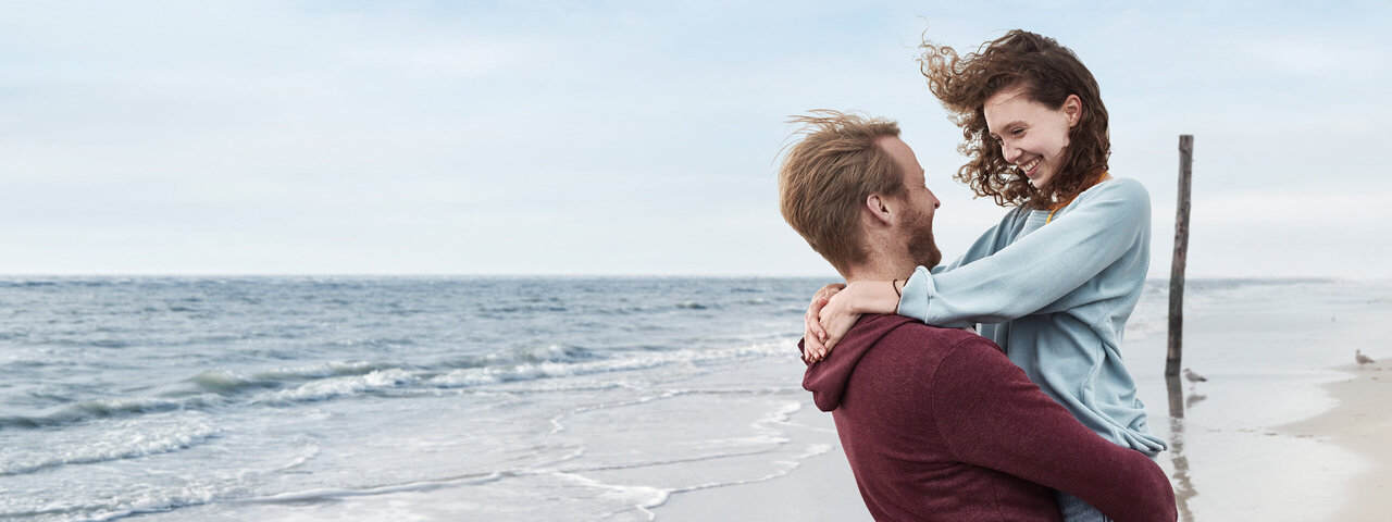 Junges Paar am Strand. Beide umarmen sich, der Mann hebt die Frau hoch und sie gucken sich innig in die Augen.