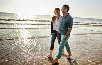  Ein Paar bei einem Spaziergang am Strand. Sie laufen barfuß durch das Wasser. Meer und Sonne im Hintergrund.