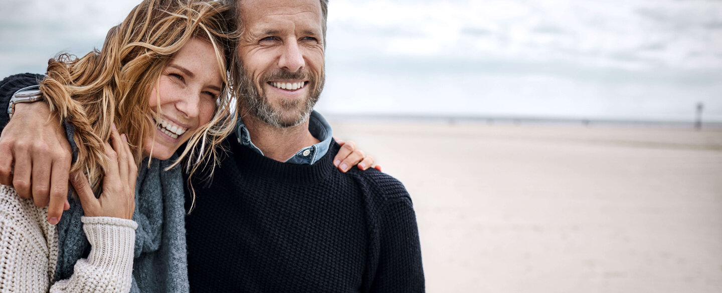 Lachendes Paar steht Arm in Arm am Strand.