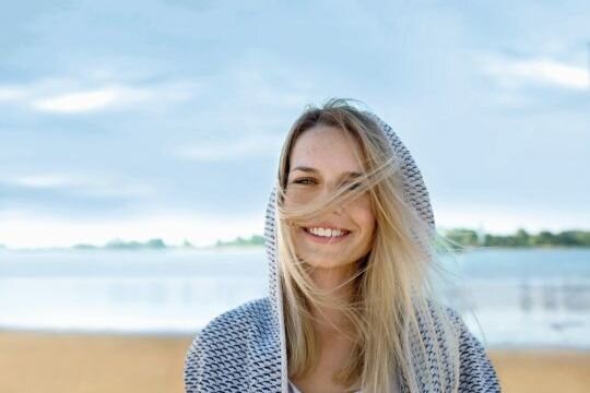 Junge Frau steht am Strand und lacht in die Kamera. Sie trägt eine Jacke mit Kapuze, die blonden Haare fliegen ihr ins Gesicht.