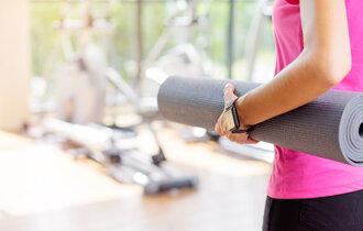 Eine Frau mit pinkem Sport-Top hält eine Yogamatte unter dem Arm und steht in einem Studio. Man sieht nur ihren Rücken.