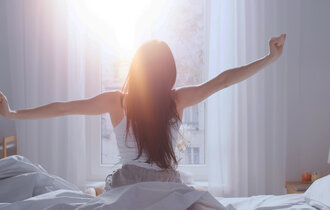 Eine Frau von hinten auf einem Bett sitzend. Sie streckt sich. Vor ihr ein Fenster, in das Licht scheint.