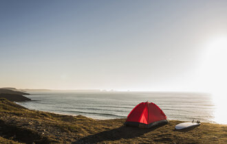 Ein rotes Zelt steht am Strand und ein Surfbrett liegt daneben während die Sonne untergeht.