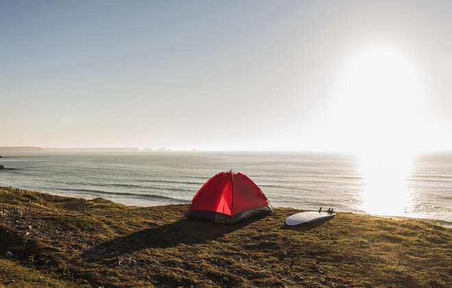 Ein rotes Zelt steht am Strand und ein Surfbrett liegt daneben während die Sonne untergeht.