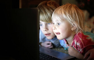 Zwei Kinder liegen in der Dunkelheit vor einem Laptop-Bildschirm, von welchem sie angestrahlt werden und gebannt darauf schauen.
