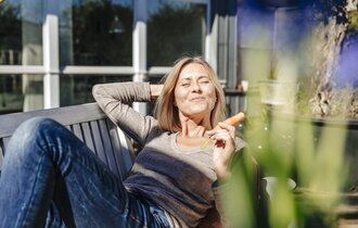 Frau legt auf einer Bank, hält eine Karotte in Ihrer Hand und hält Ihr Gesicht in die Sonne.